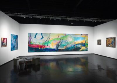Wide installation view of the PROXYCO Gallery booth at NADA Miami 2025. The monumental multi-panel painting Viendo el Monte Calvario by Lucía Vidales dominates the center wall, depicting a dreamlike landscape with floating figures, a ladder, trees, and a river in vivid blues, greens, reds, and yellows. Smaller figurative paintings hang on the left and right walls. Upholstered seating in the foreground, dark reflective floor, industrial ceiling with track lighting.