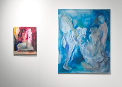 Two figurative paintings by Lucía Vidales on a white wall. A small work with warm red and pink tones depicts a seated figure. Beside it, a larger painting on a vivid blue ground shows ghostly overlapping figures rendered in pale blues, grays, and soft pinks.