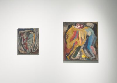 Two figurative paintings by Lucía Vidales on a white wall. A smaller square work depicts entangled forms in muted grays and reds. The larger painting beside it shows two intertwined human figures in yellow, red, and blue against a dark ground, with skulls and organic forms emerging from the composition.
