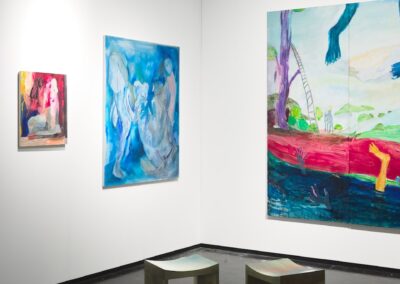 Corner installation view of the PROXYCO Gallery booth at NADA Miami 2025. The left wall features two figurative paintings by Lucía Vidales in warm and cool tones. The right wall displays a large-scale section of the multi-panel painting Viendo el Monte Calvario, showing trees, a ladder, and figures in a vivid dreamlike landscape. Two curved wooden stools with painted tops are placed in the foreground.