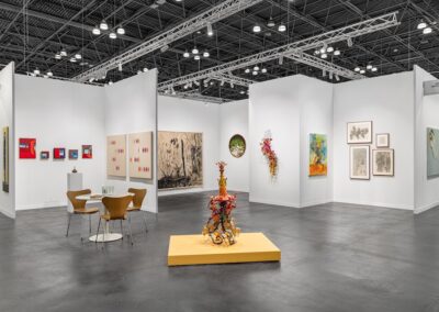 Wide installation view of the PROXYCO and Instituto de Visión joint booth at The Armory Show 2025. Works include small framed paintings on the left wall, two large beige canvases with colorful mark patterns, a large ink drawing of a forest, two circular needle-felted green textiles with red borders, a cascading wall sculpture of organic floral forms, a colorful figurative painting, and a grouping of charcoal drawings. A central floor sculpture composed of layered organic and industrial materials stands on a yellow platform. Round table and chairs in the foreground.