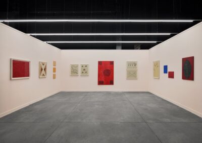 Installation view of ABRA and ProxyCO gallery booth at Feria Material 2026, featuring works by Sheroanawe Hakihiiwe. White walls display a series of paintings and drawings in deep reds and muted earth tones, depicting minimalist Yanomami iconography including geometric forms, plant and animal motifs, and symbolic figures. Concrete floor, industrial ceiling with fluorescent lighting.