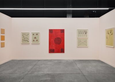 Installation view showing the left and front walls of the ABRA and ProxyCO booth at Feria Material 2026. Works by Sheroanawe Hakihiiwe include three small amber square works with dotted grid patterns, two framed drawings on pale green paper depicting a river form and geometric triangles, a large vertical red painting with a spiraling ant motif and dense border blocks at its corners, and two stacked framed works depicting radiating plant forms on pale and golden paper.