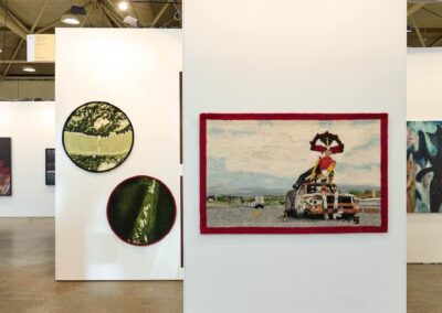 Exterior view of the PROXYCO Gallery booth at Art Toronto 2025, in the Arte Sur section. A freestanding white wall panel displays a large horizontal needle-felted textile with a red border, depicting a costumed figure atop a burned-out car on a highway. On the wall behind, two circular needle-felted works with dark borders show close-up views of green vegetation. The booth sign reading 'PROXYCO Gallery, New York' is visible in the upper right.