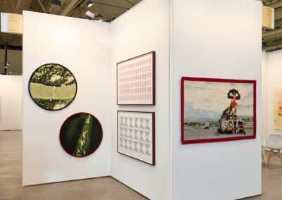Corner installation view of the PROXYCO Gallery booth at Art Toronto 2025. Left wall features two large circular needle-felted works depicting close-up views of green vegetation. Center wall displays two framed drawings — one with repeating red geometric forms, one with repeating black triangular patterns. A freestanding panel to the right holds a large horizontal needle-felted textile depicting a costumed figure atop a burned-out car.