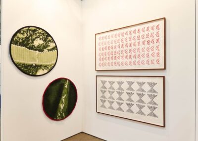 Detail of a corner wall at the PROXYCO Gallery booth at Art Toronto 2025. Two large circular needle-felted textile works hang on the left — one with a dark border showing a forest canopy, one with a red border showing a close-up of a green surface. Two framed drawings on the right wall display repeating geometric patterns, one in red and one in black on white paper.