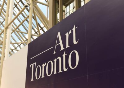 Entrance signage for Art Toronto 2025. The fair's name is displayed in large cream-colored serif type against a deep purple background panel. A glass atrium ceiling and steel structural beams are visible above.