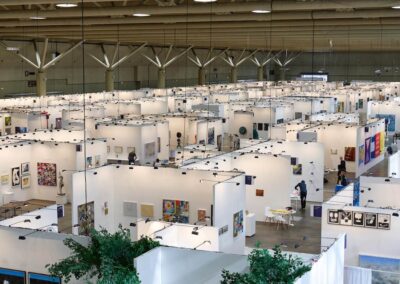 Aerial overview of the Art Toronto 2025 fair floor, photographed from an elevated vantage point through glass. Rows of white booth walls extend across a large convention hall, with colorful artworks visible throughout. Visitors and gallery staff are seen among the booths. Potted trees are visible in the foreground.