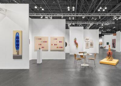 Exterior view of the PROXYCO and Instituto de Visión booth at The Armory Show 2025, booth 135. A tall narrow blue figurative painting is visible at the far left alongside a small ceramic sculpture on a pedestal. Two large beige canvases with colorful marks, the cascading wall sculpture, a grouping of framed drawings, and the central floor sculpture on a yellow platform are visible within the booth.