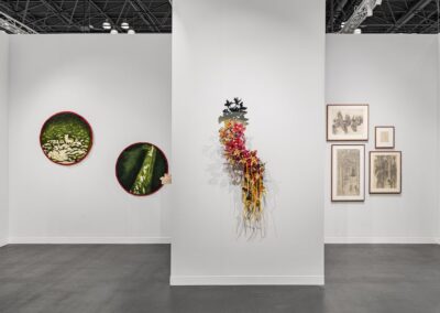 Detail view of three wall sections in the PROXYCO and Instituto de Visión booth at The Armory Show 2025. Left wall displays two large circular needle-felted green textile works with red borders. Center wall features a cascading wall sculpture of layered flowers and organic forms in reds, yellows, and black. Right wall shows four framed charcoal drawings arranged in a cluster.