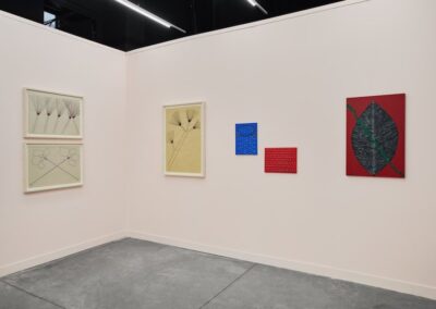 Corner installation view of the ABRA and ProxyCO booth at Feria Material 2026. Left wall displays two stacked framed works depicting radiating plant forms and circular floral figures. Center wall shows a large framed botanical drawing on golden paper, two small works on vibrant blue and red grounds, and a large painting of a dark leaf form on deep red with a green vine.