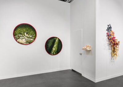 Corner detail of the PROXYCO and Instituto de Visión booth at The Armory Show 2025. Two circular needle-felted green textile works with red borders hang on the left wall, one depicting a small animal among stones, the other a close-up of a green surface. A small ceramic shelf piece and a partial view of the cascading floral wall sculpture are visible on the adjacent wall.