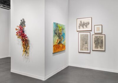 Corner installation view of the PROXYCO and Instituto de Visión booth at The Armory Show 2025. A cascading wall sculpture of layered organic floral forms in reds and yellows hangs on the left wall. Center wall features a large colorful painting of a swirling tree form over an orange ground with a small red figure. Right wall displays four framed charcoal drawings in a gallery arrangement.