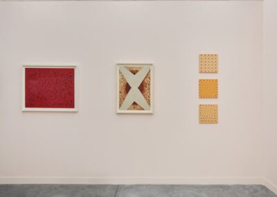 Detail view of a single wall in the ABRA and ProxyCO booth at Feria Material 2026. Three works by Sheroanawe Hakihiiwe are shown: a red painting with scattered dark mark-like forms, a large framed X-cross composition on aged natural fiber, and three small vertically stacked amber square works with dotted border patterns.