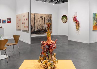 View from inside the PROXYCO and Instituto de Visión booth at The Armory Show 2025, looking toward the central floor sculpture on a yellow platform. In the background, booth walls display two beige canvases with colorful marks, a large ink drawing of a forest, a circular green textile, a cascading floral wall sculpture, and a colorful figurative painting. Chairs and a side table are visible at left.