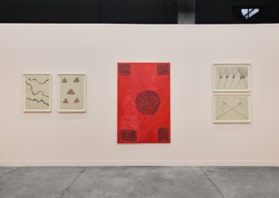Front wall installation view of the ABRA and ProxyCO booth at Feria Material 2026. Two framed drawings on pale paper depict a sinuous river form and geometric mountain triangles. A large vertical red painting dominates the center, featuring a dense spiraling ant motif surrounded by dark border blocks at each corner. Two stacked framed works on the right depict radiating plant forms and circular crossed botanical figures.